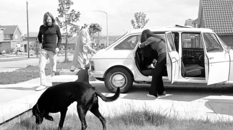 Getty Images Lennon stands by a white car while Ono leans into the vehicle. A black dog can be seen on a patch of grass in front of the car while another person stands by the boot. A row of houses can be seen in the background.