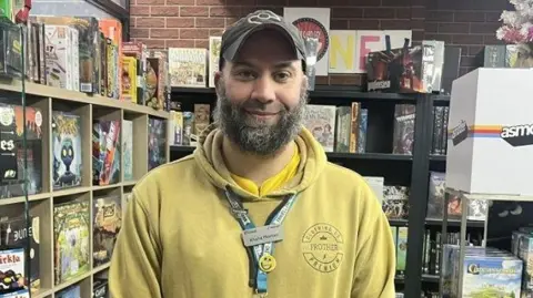 Rachel Russell/BBC Khalid Morton wearing a yellow jumper with his work lanyard on and also a brown cap. He is smiling and has a dark beard with flecks of grey in it. He is stood in front of shelves in the shop that are filled with old comics, novels and board games.