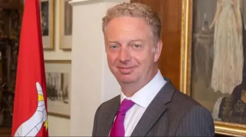 Alex Allinson smiling at the camera. He has fair hair and is wearing a grey suit, white shirt and pink tie, and is standing next to a red Manx flag.