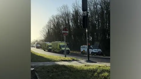 A road junction with two ambulances parked on the side of the road. There is a set of traffic lights and a pedestrian crossing. In the background you can see the front of a red bus that is parked on the verge.