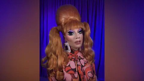 Supplied A drag queen wearing purple eyeliner and bright red hair with long pigtails. She is wearing a bright pink and orange dress.
