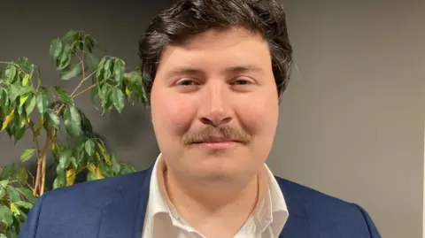 A close up of Deputy Tom Rylatt. He has short black hair, a brown moustache and is wearing a white shirt and navy jacket.