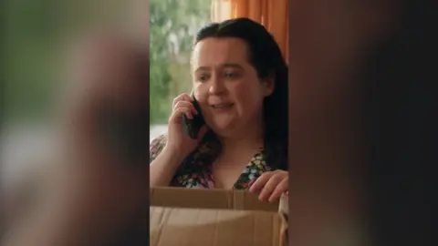 A woman, Nina, is on the phone. She stands with her hand on a box.