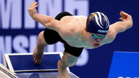 PA Bruce Dee wearing black swimming trunks, goggles and a swimming cap, dives into a pool 