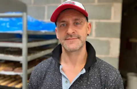 Baker Simon Hill wearing a cap standing in front of shelves of bread 