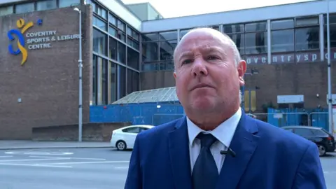 BBC Councillor Jim O'Boyle outside the former site of 'Cov Baths'. He is wearing a blue suit and tie, with a white shirt and is standing across the road from the building, where a sign saying Coventry Sports and Leisure Centre can be seen.