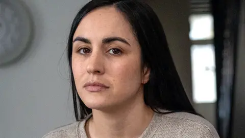 A woman with dark hair and a grey top looks into the camera.