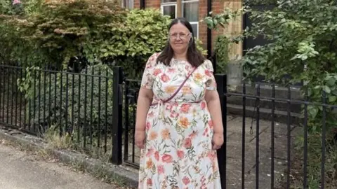 A woman with long dark hair stands in front of black railings with a brick building just visible behind them. She is wearing glasses and a long white dress with short sleeves and a pink and yellow flower pattern all over.