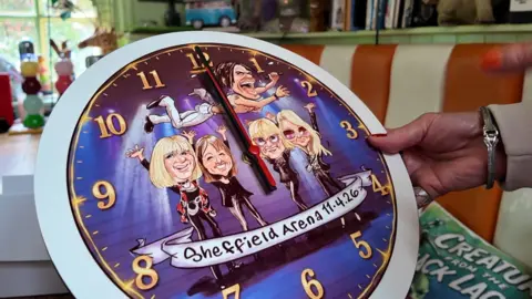 A clock with the purple face designed as a caricature of four blonde women and a man flying over their heads