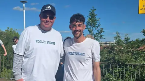 Wigmore Primary School Mr Marshall on the left and Mr Whadcoat on the right. They are both wearing white T-shirts with "Wembley 2 Wigmore" on the chest and are smiling. They are standing at the school. 