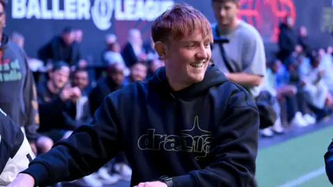 Angryginge at a Baller League event. The streamer has red hair with a long fringe and smiles while looking over his left shoulder. He wears a black hoodie and in the background, rows of people sit pitchside. 