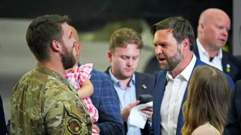 A man in military uniform is holding a young child in a pink, gingham dress in his arms. He is talking to JD Vance, who is wearing a white shirt with no tie and a blue jacket. A woman appears to be watching on, with the back of her head and her long, brown hair visible. Security personnel can be seen in the background.
