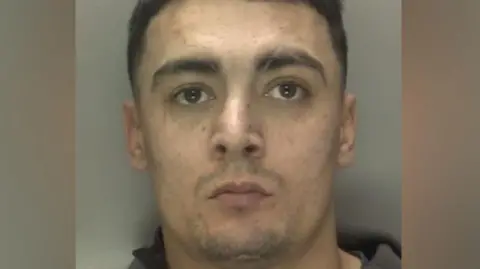 A police mugshot shows a man with short brown hair and light stubble. He is wearing a dark grey top.