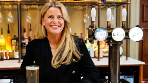 British Beer and Pub Association Emma McClarkin, standing in a pub, with a drink in front of her. She is smiling, with long blonde hair and hoop earrings in her ears. She is wearing a black top with gold buttons. 