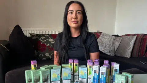 Cara is a 25-year-old woman with long dark hair, she is wearing a black T-shirt and she is sitting on a sofa. On a table in front of her is an array of multi-coloured disposable vape boxes.