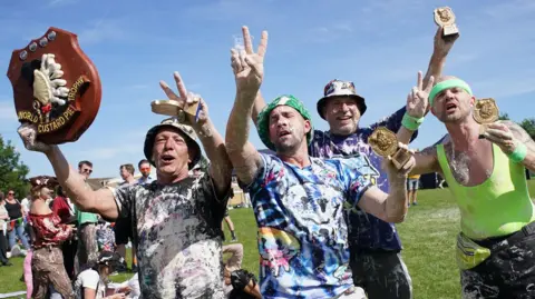 PA Media Several people, covered in custard pie, are celebrating after winning a competition. One of them is holding a trophy which reads "world custard pie trophy".