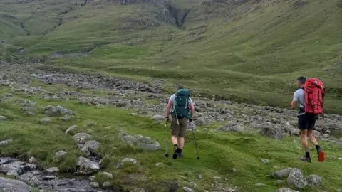 Two men walking along a flat expanse of grass with lots of rocks. They are both wearing t-shirts and shorts and the man on the right has a red walking backpack and the one on the left has a green and is using two walking poles. The ground is steep all around them.