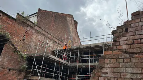 A worker climbs a ladder among the ruins of the theatre. He is surrounded by scaffolding and bomb-damaged brick work.