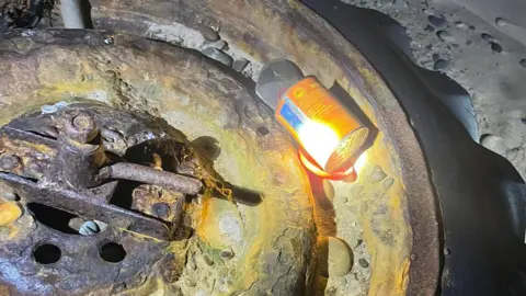 The flare is in an orange can which appears partly opened. It lies on the inside of a rusted tyre, which is lying on the beach.