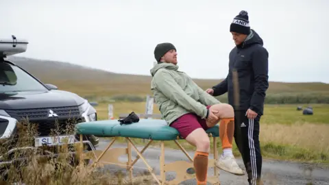  Robbie Guthrie/PA Seb Key on a massage bed feeling his leg, with a member of his team watching over him. They are on the side of a road.