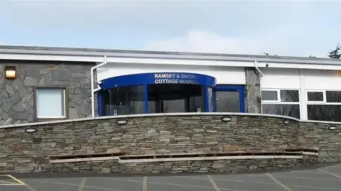 The front entrance to Ramsey Cottage Hospital, which has a blue revolving door with a stone wall in front of it and a car park.