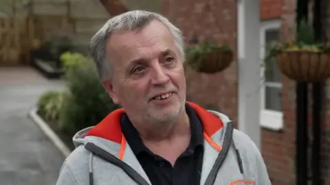 John Frosdick, who has white hair and a neat beard, can be seen wearing a grey hooded jacked over a black polo shirt as he stands outside his home. 