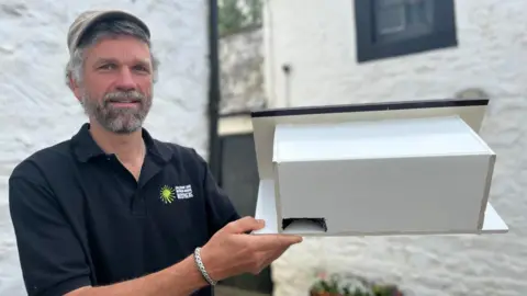 A man with grey hair and a beard in a cap and black T-shirt holds up a white bird box