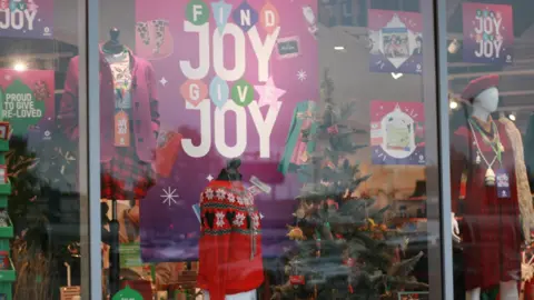 The window of a charity shop with various Christmas items and clothes on display such as festive sweaters and a Christmas tree. There are placards reading "Find Joy, Give Joy" behind the mannequins.