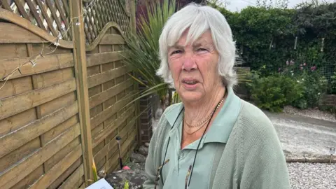 Jennie Warren looks cross while standing next to her garden fence. She is wearing a green shirt and green cardigan.