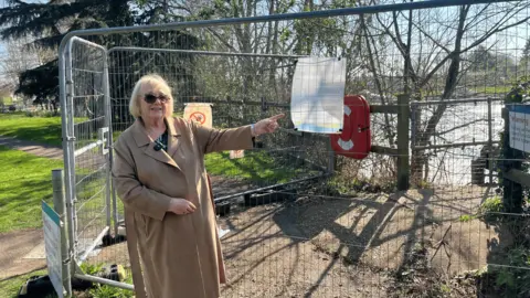 Blonde woman wearing sunglasses and brown coat next to fence and pointing at notice on it
