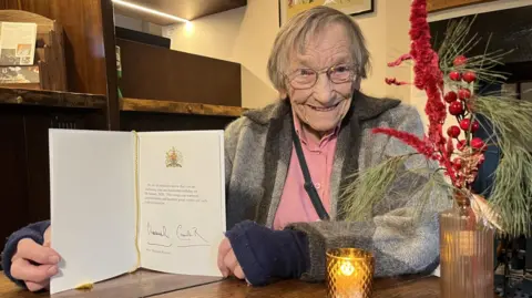 Dr Margaret Bradshaw is in a grey knitted jumper with her 100th birthday card from the King and Queen held open in her hands. A candle is next to her alongside a small vase containing red flowers. She has short grey hair and glasses.