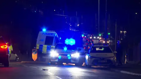 Emergency service vehicles including a police van and cars on Bolton Road in Ashton-in -Makerfield in front of a line of traffic. It is dark.