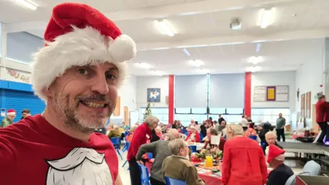 Company at Christmas Man dresses in a santa hat taking selfie with people eating in background.