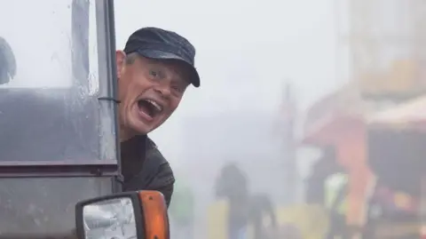 Martin Clunes laughs, mouth wide, towards the camera as he leans out the side of a utility vehicle. It's a wet, rainy day. In the background in the mist is a colourful traditional funfair. Clunes is wearing a black baseball cap and a black coat.