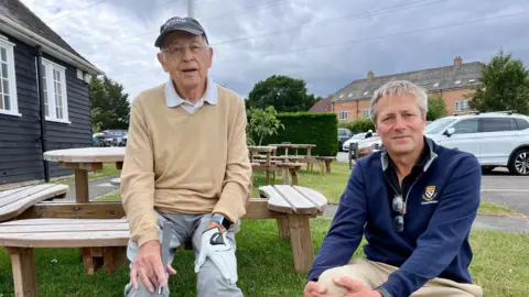 An elderly man sitting on a picnic bench holding a golf club while looking into the camera. Sat on a patch of grass beside him is a man, also looking into the camera. Behind them are more benches, a small black building and a car park
