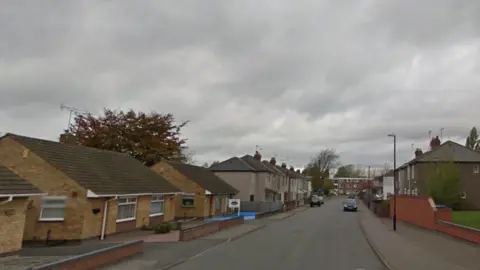 Houses and bungalows line the road with some cars parked on either side of the street.