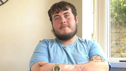 Will, who has short brown hair and wears a blue t shirt, looks at the camera with his arms folded and a neutral expression