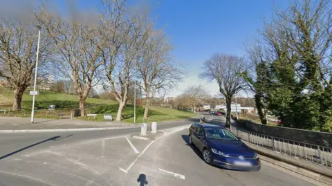 A blue car leaving Kings Road junction with has a large field to the left of it and trees either side. 