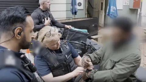Three immigration officers putting handcuffs on a man. He has dark skin and is wearing a green jacket. There is an e-bike behind him. His face has been blurred so we do not know who he is. 