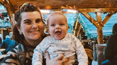 Tyler O'Brien holds her young son, Alfie, as they sit in a Christmas market. They are both smiling.