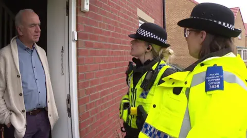 Two female police officers, dressed in uniform, talk to a man standing at his front door wearing a cream cardigan and blue shirt 