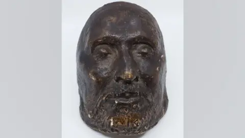 The Cromwell Museum A funeral mask for Oliver Cromwell. It shows his bearded face. His eyes are closed. It is made from a very dark material