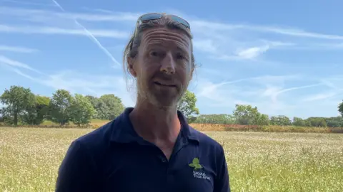 A man in a blue polo shirt pictured in a dry grassy field. He has sunglasses on his head and is smiling. 
