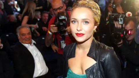 Hayden Panettiere surrounded by photographers she has her hair tied back and wearing red lipstick and a greendress with a black leather jacket over the top