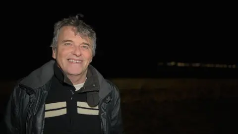 Dave smiles at the camera as the light shines on his face. He is wearing a black leather jacket with a black zip-up jumper underneath which has four white marks on He has short grey hair and his hands are held behind his back.