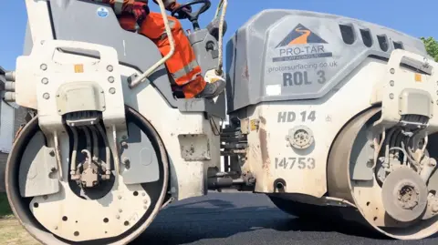 London Luton Airport A close up of a worker operating a roller machine to smooth recycled asphalt from London Luton airport. He is wearing orange overalls. 
