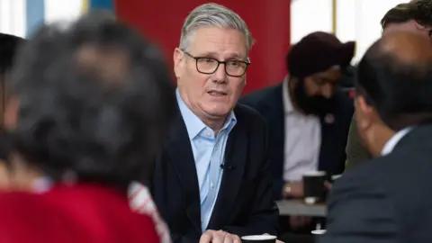 PA Media Sir Keir Starmer - who has grey hair, glasses, a blue shirt and black jacket - talks to people watching him as he sits at a table.