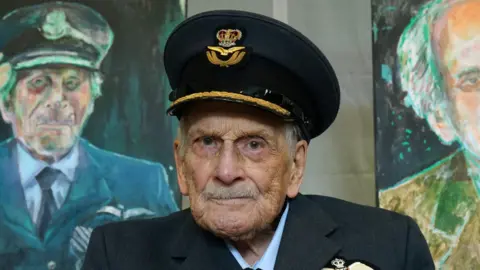 Group Captain John 'Paddy' Hemingway sits in front of two painted portraits and looks at the camera. He is wearing an Air Force military hat and uniform.