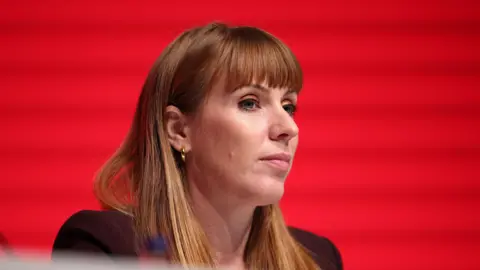 Deputy Prime Minister, Angela Rayner, at the Labour Party Conference in Liverpool, Britain, seen wearing a mauve blazer against a red background. 