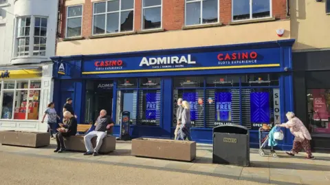 A gaming centre shop front which is bright blue with red and white lettering. There are seats outside with a man and woman sitting down. A woman pushes a mobility scooter behind them. 